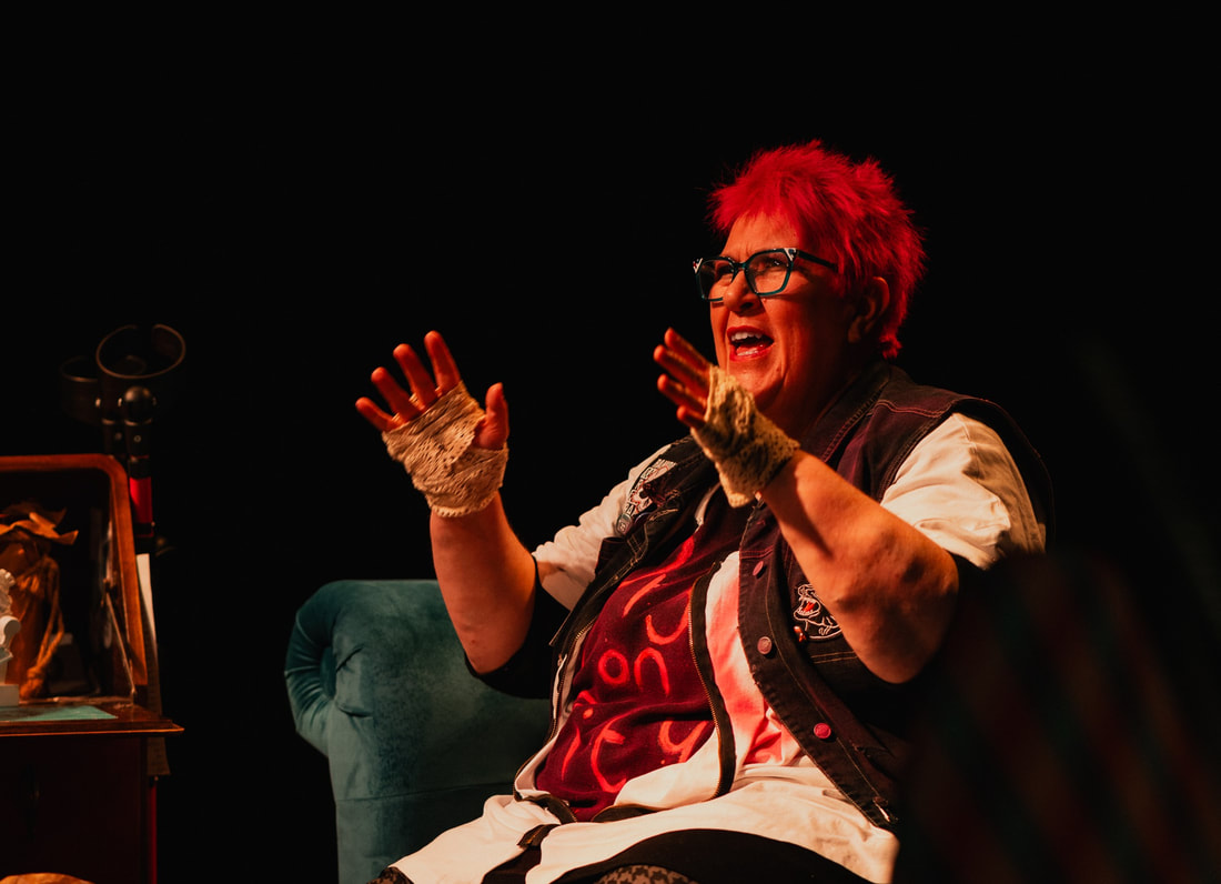 Actor and writer Vici Wreford-Sinnott is an older white woman with pink, purple and blue hair. She is shouting/laughing as she tears through a huge piece of crumpled brown parcel paper. The word UNRULY is written on the paper. 