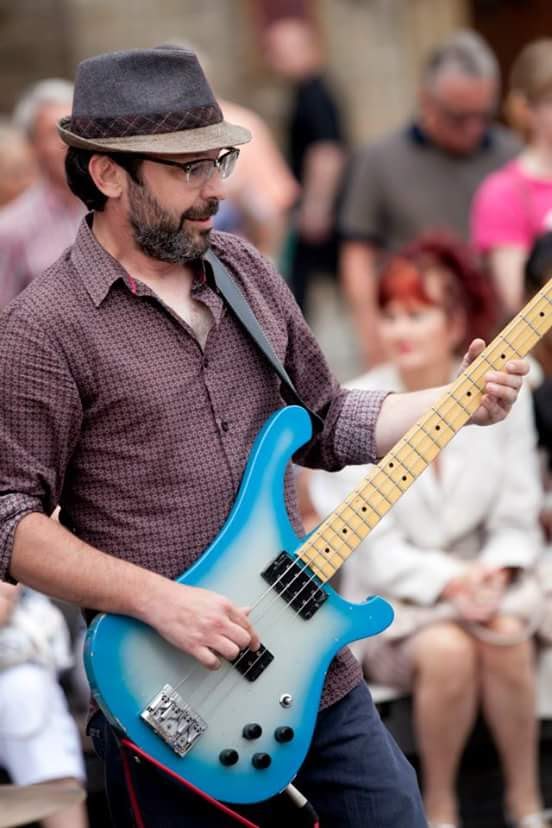 An older white man, playing guitar at an outdoor festival. He has a beard and a pork pie hat.  
